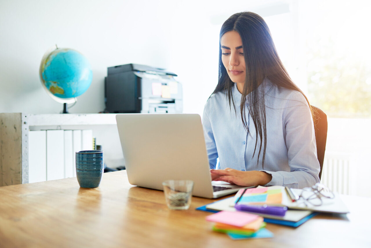 Competent young business secretary working at a laptop at a wooden desk surrounded by colorful memo notes to organise and prioritise her tasks